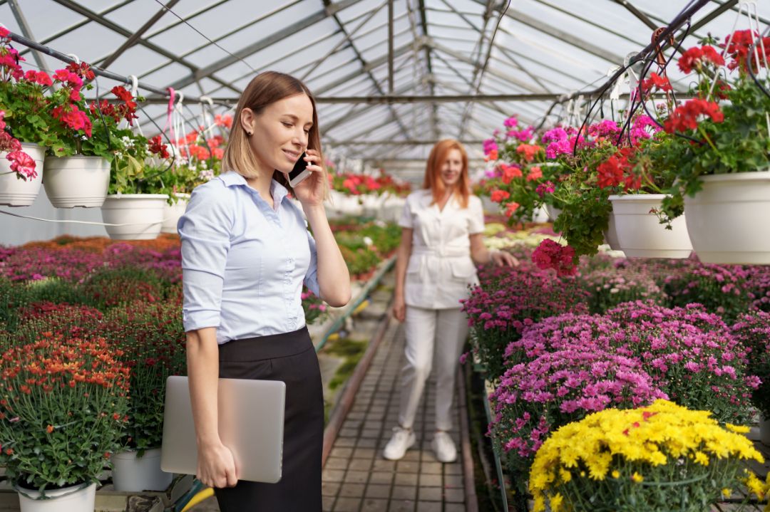 Businesswoman discussing on the phone a proposal. She holds a laptop in a green house with flowers. Small business development concept.