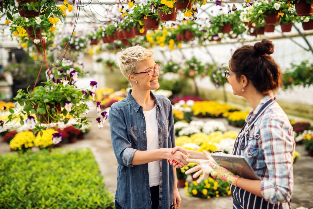 Female florist dressed in apron with tablet in hands shaking hands with customer while standing in greenhouse.