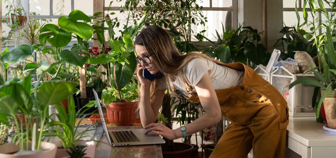 Busy entrepreneur woman, female gardener work on laptop, order plants by phone in home garden. Young businesswoman in jumpsuit check document on computer, speak on mobile in office full of houseplants