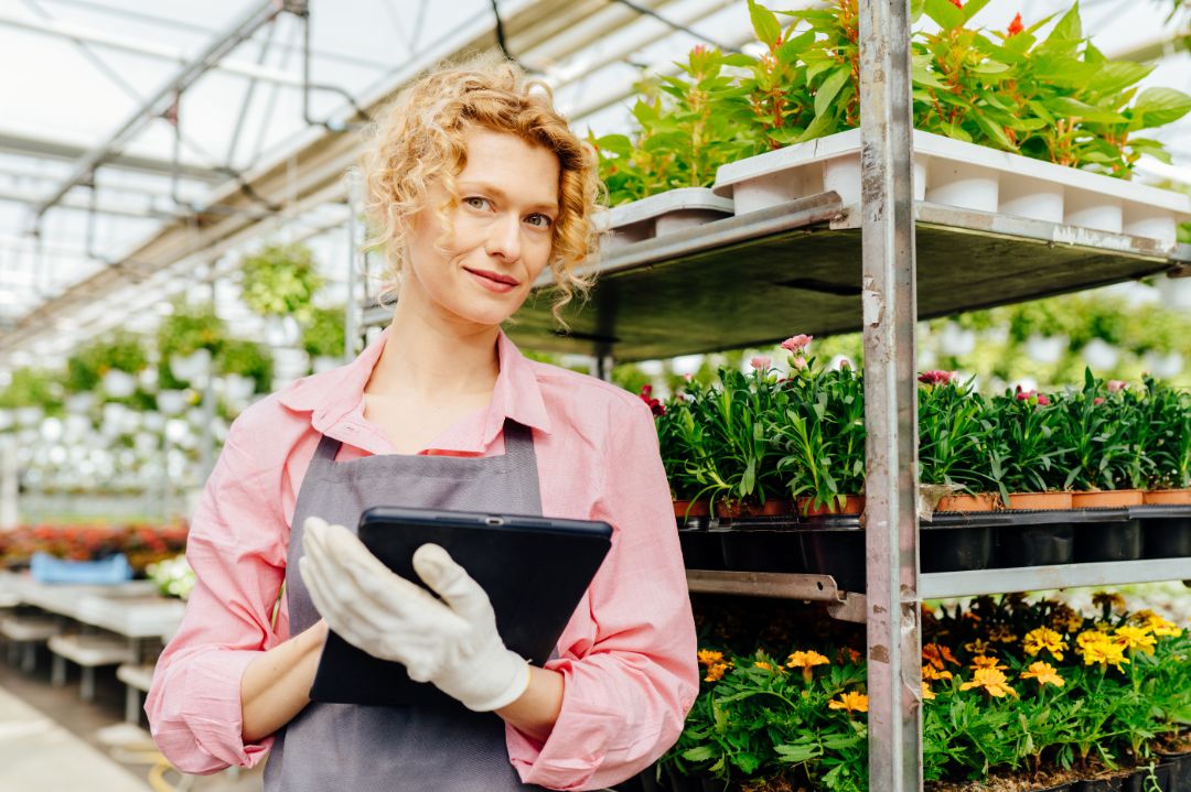 Portrait of attractive woman gardener in grey apron and gloves working with tablet and everyday taking care of the plants in big industrial greenhouse center. Gardening, profession and people concept.