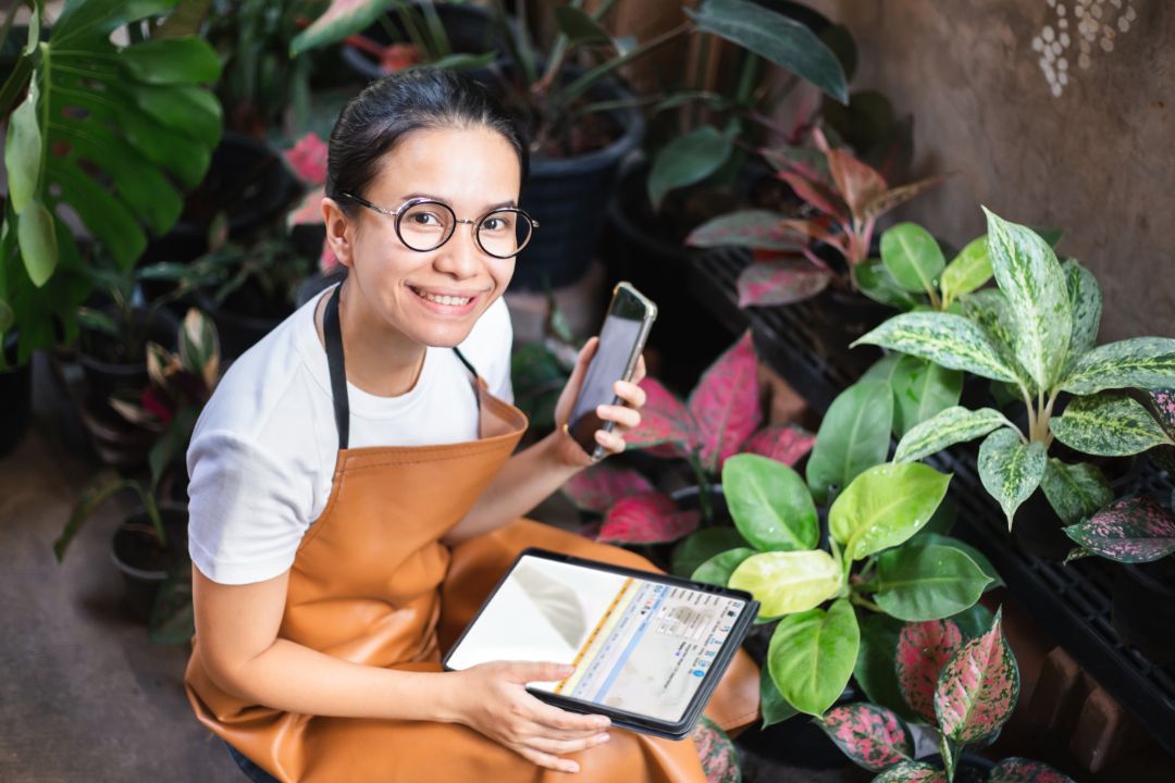 Asian gardener woman working with smartphone or tablet and looking at camera in tree shop. Smiling  female using device technology for sale houseplants to customer. Home business owner
