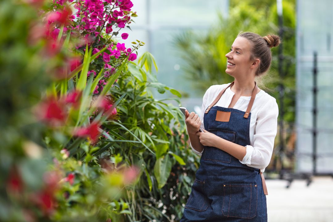 Modern technology in gardening business. Portrait of female environmentalist using digital tablet in greenhouse. Confident female is wearing apron while working. She is standing by flowering plants